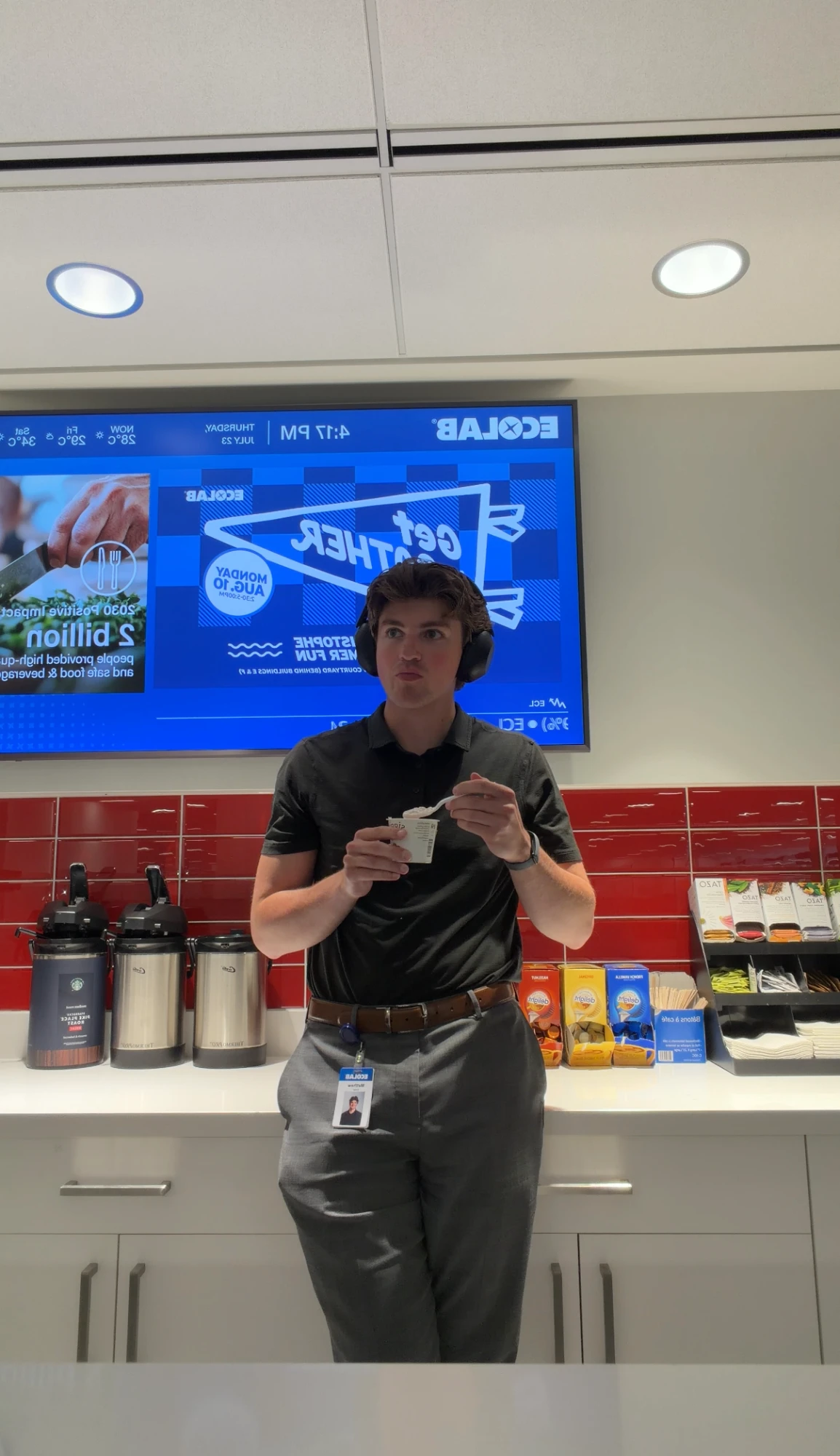 Eating from a cup in the office kitchen, wearing headphones and a work badge